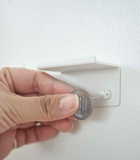 A hand is shown using a coin to secure nails into a wall, installing a white wall-mounted phone/tablet holder. view 7