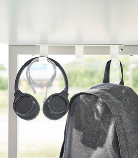Three matte white metal hooks are secured to the underside of a table. One hook holds a pair of over-ear headphones and a wrapped charger cord, while another holds a backpack. view 5