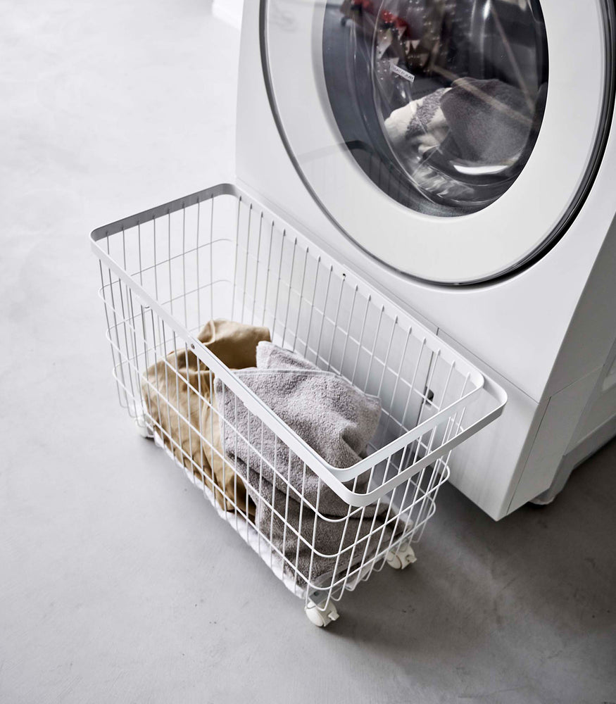 View 5 - Rolling Wire Basket by Yamazaki Home in white in front of a dryier with several towels inside.