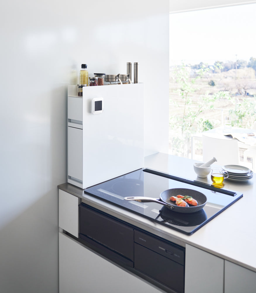 View 3 - White Yamazaki Home Two-Tier Sliding Drawer Seasoning Rack next to a stove top with oil and spices stored