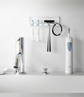 Yamazaki Home's white Traceless Adhesive Toothbrush Holder on a bathroom wall, holding two toothbrushes and a razor beside a faucet. view 4