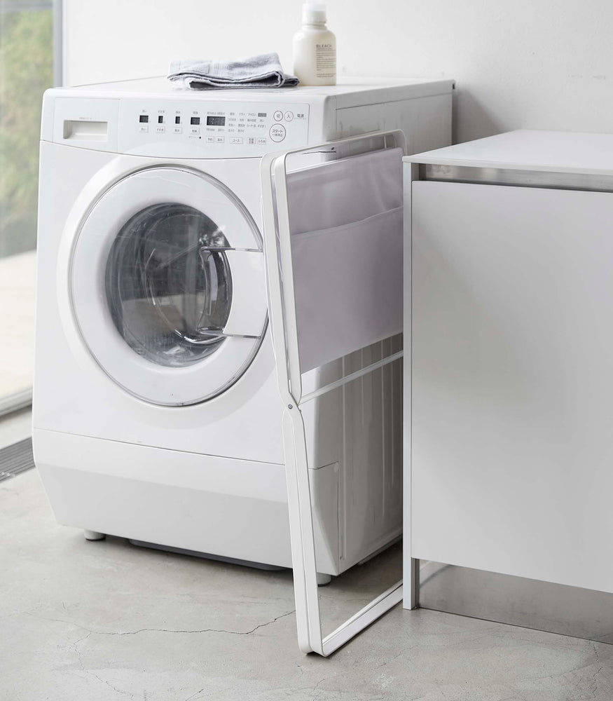 View 5 - A white canvas hamper with white metal legs is folded up and leaned between a washing machine and table. A towel is folded on top of the washing machine.