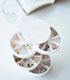A bird’s-eye view of a white four-tier swivel accessory holder on a white surface. The accessory holder’s tiers and lid are swiveled opened so the inside contents can be seen, each tier has 7 individual compartments in a pie-shaped fashion. Each individual compartment of the tiers holds various beads and jewelry-making parts. In the background are a clear glass cup and an opened book. view 14