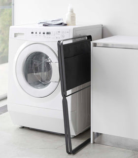 A black canvas hamper with black metal legs is folded up and leaned between a washing machine and table. A towel is folded on top of the washing machine. view 25