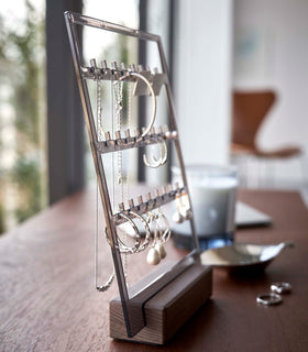 A profile view of an acrylic translucent mauve earring holder with a rectangular wooden base on a dark wood dresser. The acrylic holder has upward pointed hooks and slots placed in an interchangeable pattern. Hanging from the hooks are chained necklaces, and in the slots are various earrings. Out-of-focus behind the product are two rings and a leaf shaped catch-all dish. view 13