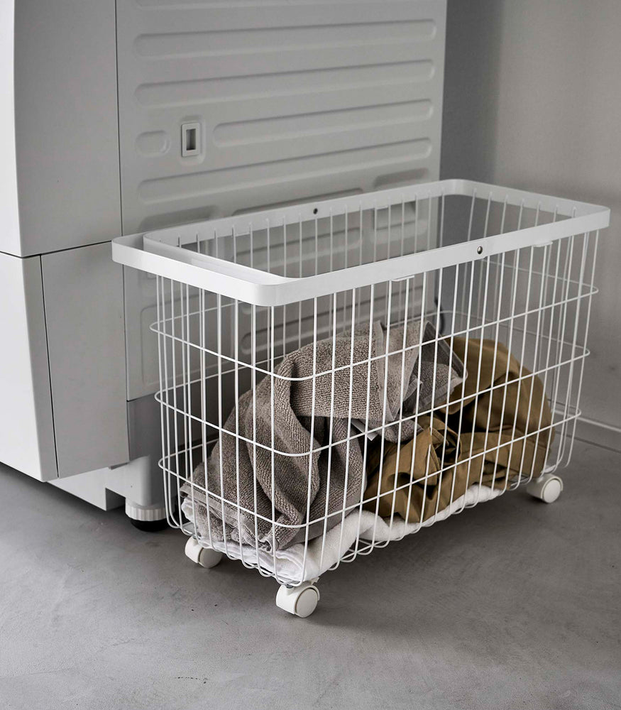 View 6 - Rolling Wire Basket by Yamazaki Home in white stowed next to a laundry machine with several towels inside.
