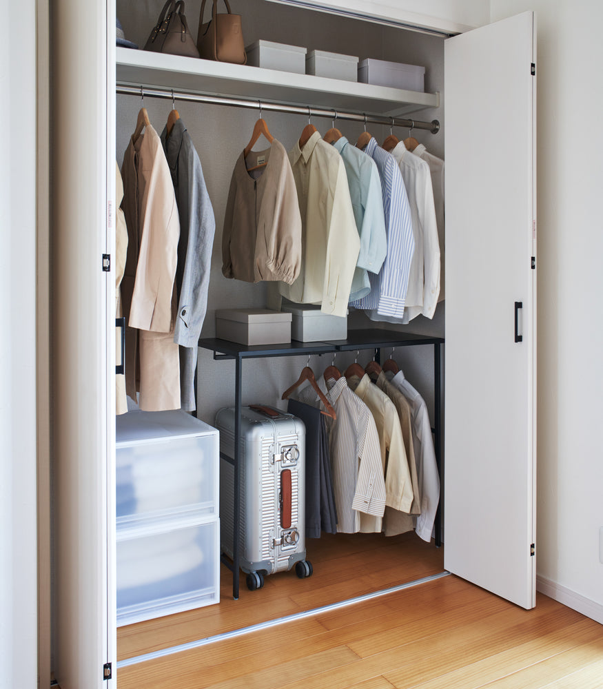 View 15 - Black Yamazaki Home Expandable Suitcase Rack in a closet with clothes hung and a suitcase underneath