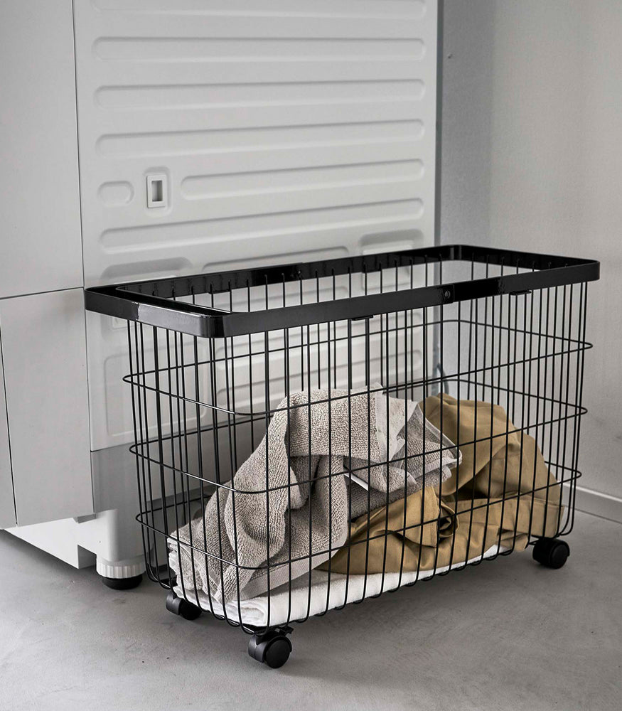 View 14 - Rolling Wire Basket by Yamazaki Home in black stowed next to a laundry machine with several towels inside.