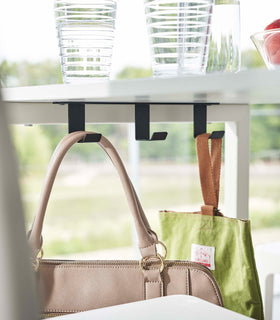 Three matte black metal hooks are firmly attached to the underside of a small white dinner table. Two of the hooks securely hold onto purses. view 8
