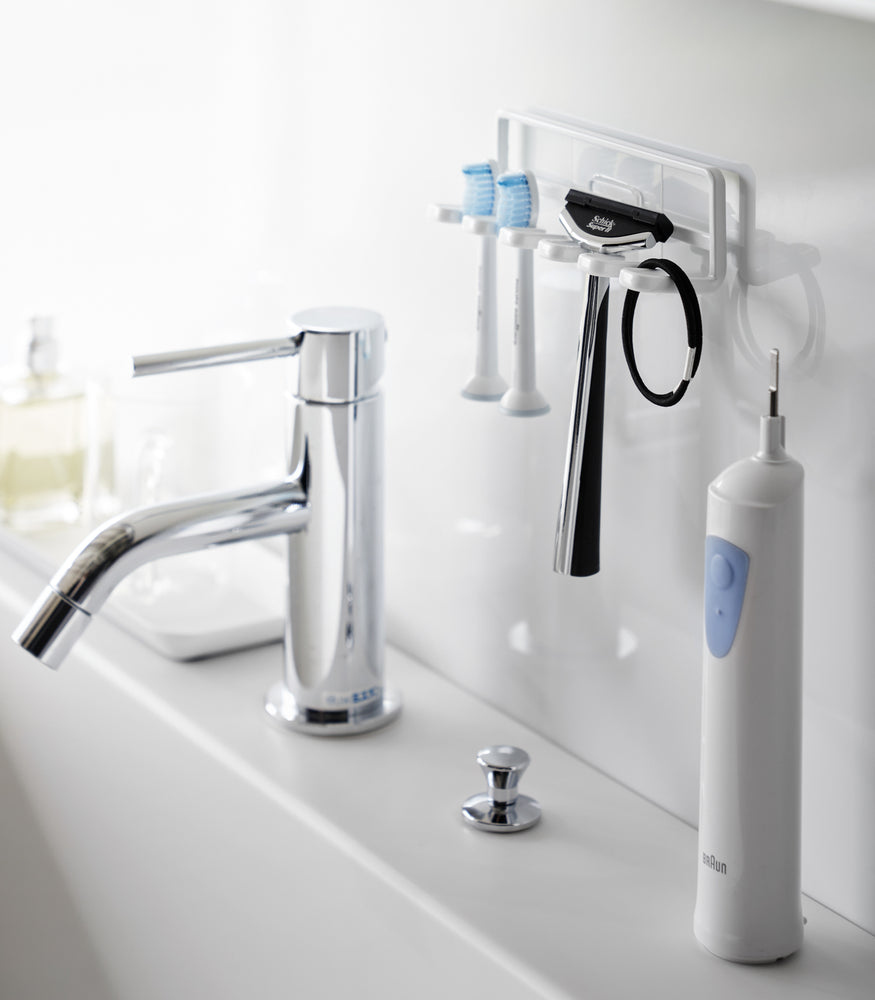 View 3 - Yamazaki Home's white Traceless Adhesive Toothbrush Holder on a bathroom wall, holding two toothbrushes and a razor beside a faucet.