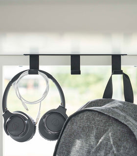 Three matte black metal hooks are secured to the underside of white desk. One hook holds a pair of over-ear headphones and a wrapped charger cord, while another holds a backpack. view 10