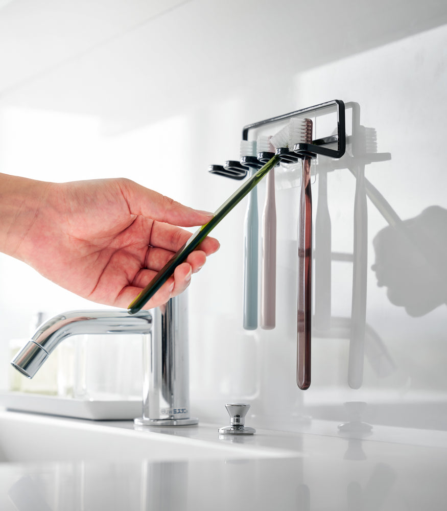 View 10 - Yamazaki Home's Traceless Adhesive Toothbrush Holder, black, mounted on a bathroom wall with four colorful toothbrushes and a chrome faucet.