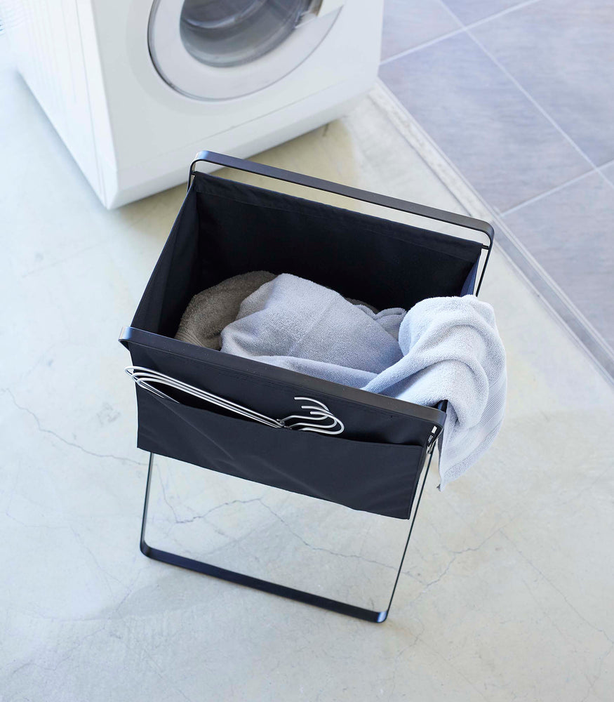 View 15 - Bird’s eye view of a black canvas hamper with multiple towels inside and wired hangers peeking out of a side-pocket. A washing machine is seen in the background.