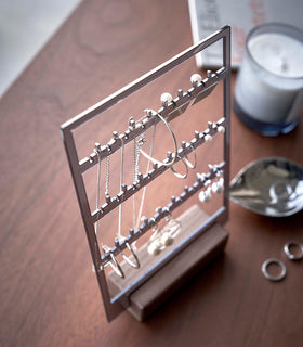 A birds-eye view of an acrylic translucent mauve earring holder with a rectangular wooden base on a dark wood dresser. The acrylic holder has upward pointed hooks and slots placed in an interchangeable pattern. Hanging from the hooks are chained necklaces, and in the slots are various earrings. On the surface of the dresser, in front of the earring holder, are two rings next to a decorative catch-all plate. view 10