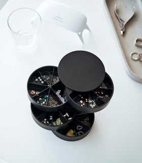 A bird’s-eye view of a black four-tier swivel accessory holder on a white surface. The accessory holder’s tiers and lid are swiveled opened so the inside contents can be seen, each tier is divided into seven individual compartments in a pie-shaped fashion. Each individual compartment of the tiers holds various beads and jewelry-making parts. In the background is an oval shaped catch-all plate with a pair of silver needle nose pliers and three rings. view 22