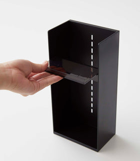 A male hand pulls a black transparent tray to adjust the location in a cosmetics organizer. It is a black resin rectangular cosmetics holder with an open face and top. The removable tray acts as a shelf and has an upward facing lip along the edge to prevent products from falling out. view 17