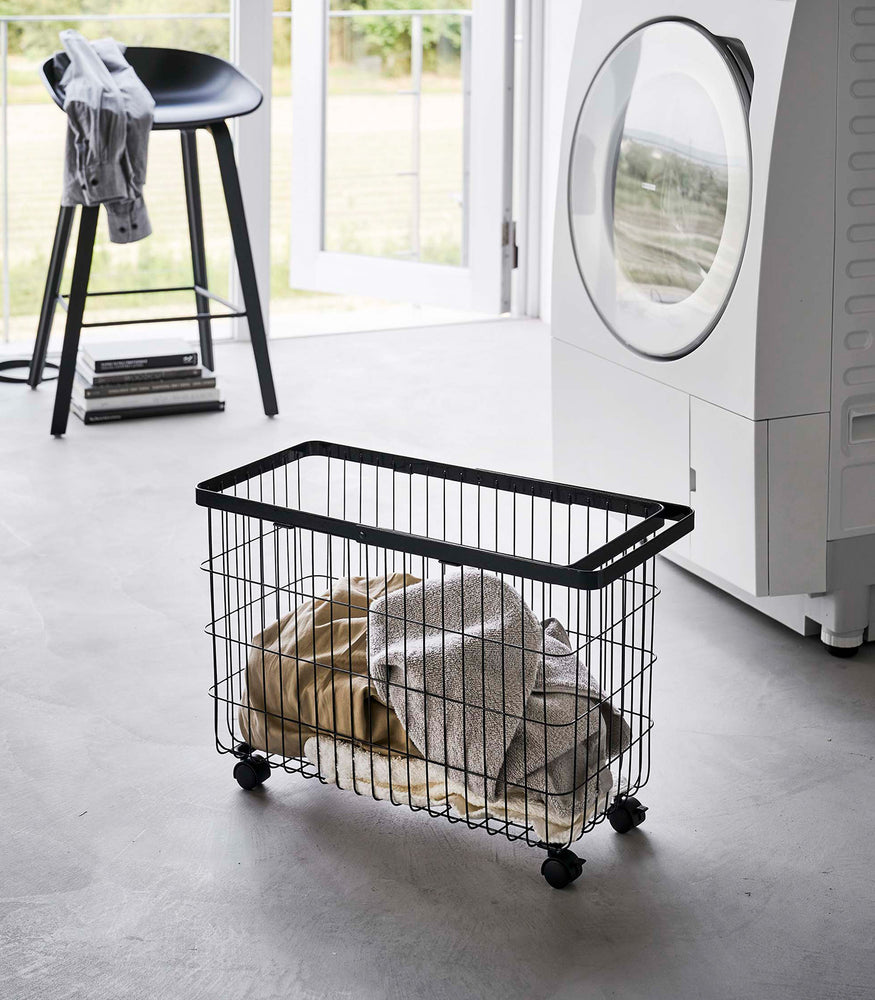 View 10 - Rolling Wire Basket by Yamazaki Home in black in a sunny laundry room with several towels inside.