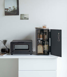 A vertical black metal breadbox is seen on a white kitchen counter next to a black microwave oven and a glass drip coffee pot with a dark liquid inside. view 22