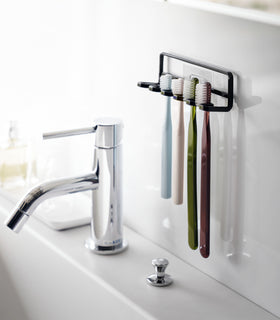 Yamazaki Home's Traceless Adhesive Toothbrush Holder, black, mounted on a bathroom wall with four colorful toothbrushes and a chrome faucet. view 7