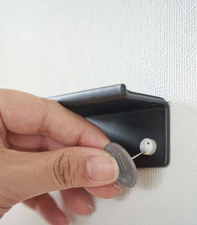 A hand is shown using a coin to secure nails into a wall, installing a black wall-mounted phone/tablet holder. view 14