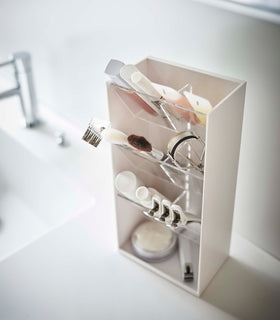 An angled view of a white rectangular resin cosmetics organizer on a white bathroom counter. It has an open face and top and three deep transparent trays that sit diagonally with adjustable transparent dividers placed in the middle of each tray for easy visibility. view 22
