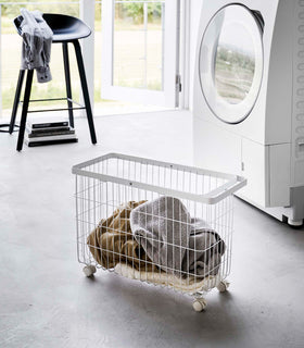 Rolling Wire Basket by Yamazaki Home in white in a sunny laundry room with several towels inside. view 2