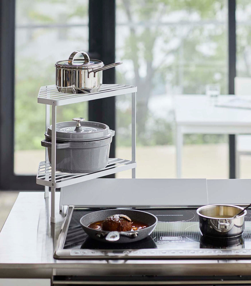 View 3 - Front view of Black Two-Tier Corner Riser holding pot and saucepan next to stovetop by Yamazaki Home.