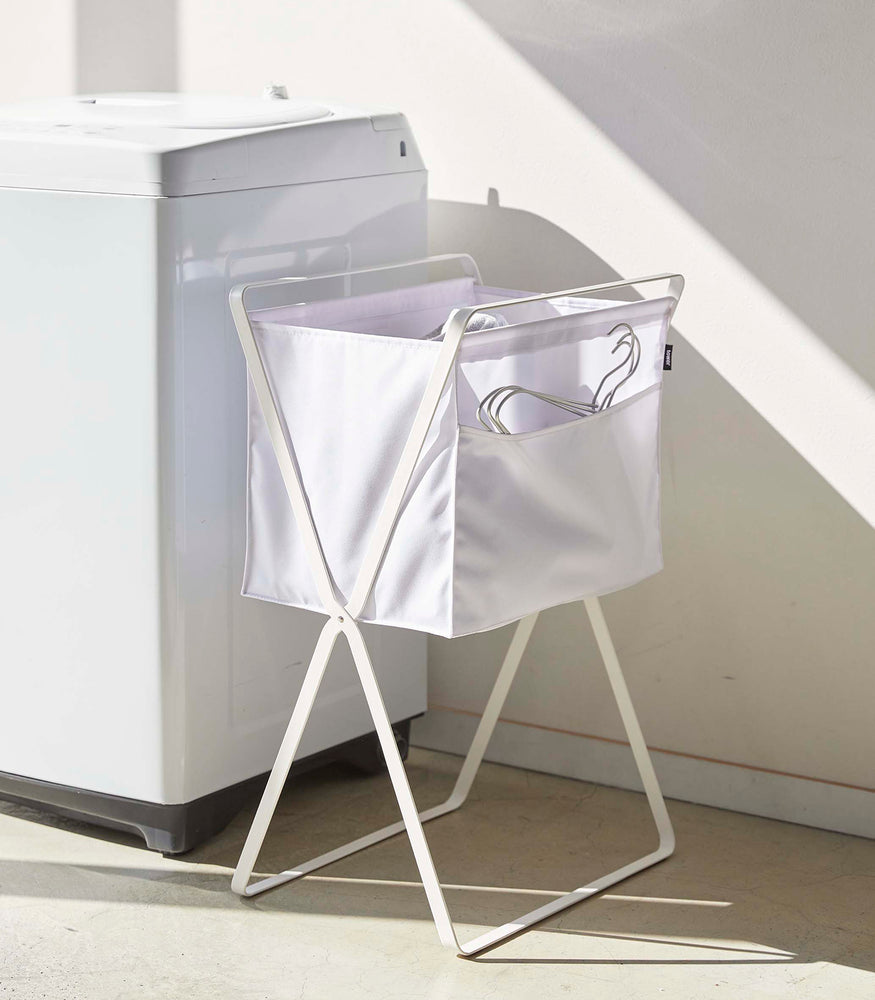 View 3 - A white canvas hamper with white metal legs is positioned next to a washing machine. Wired hangers are seen poking out of a side pocket on the hamper.