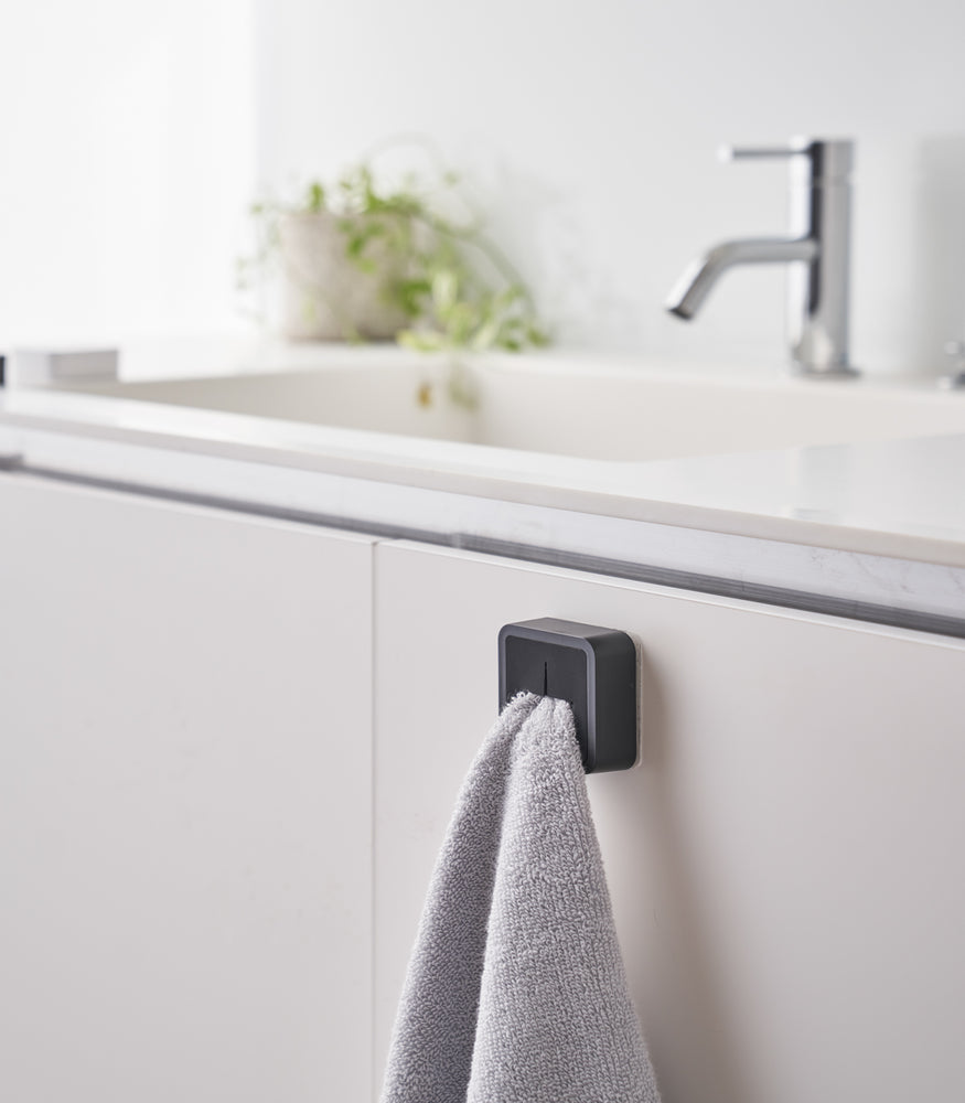 View 15 - Black Yamazaki Home Traceless Adhesive Towel Holder attached to a cabinet under a sink with a towel inserted