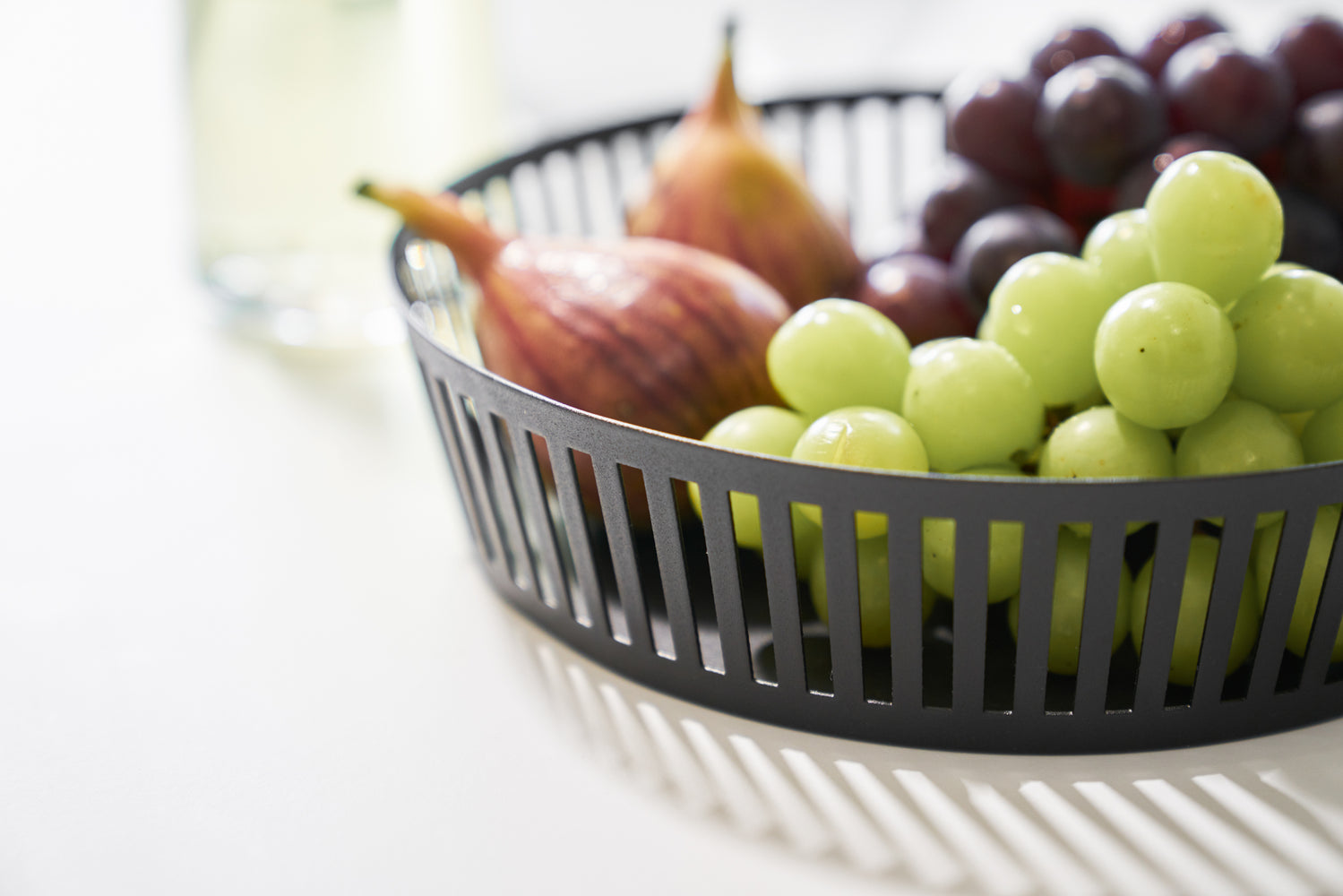 View 8 - Close up view of black Fruit Basket holding grapes and figs on white tabletop by Yamazaki Home.