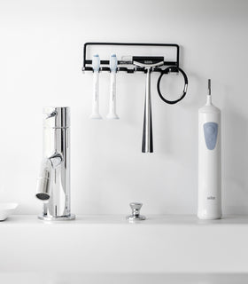 Yamazaki Home's black Traceless Adhesive Toothbrush Holder on a bathroom wall, holding two toothbrushes and a razor beside a faucet. view 9