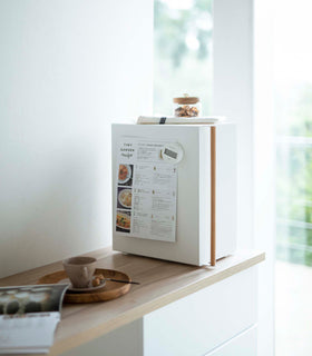 A profile view of a vertical white metal bread box sat on a wooden kitchen counter.  A long vertical wooden handle is positioned on the left at the front of the box. A magnetic timer is holding up a recipe card on the side of the breadbox. A folded towel and glass container of treats is laid on top. On the counter in next to the breadbox is a wooden plate with a brown coffee cup and matching saucer almost out of focus. Next to the wooden plate is an opened magazine. view 12