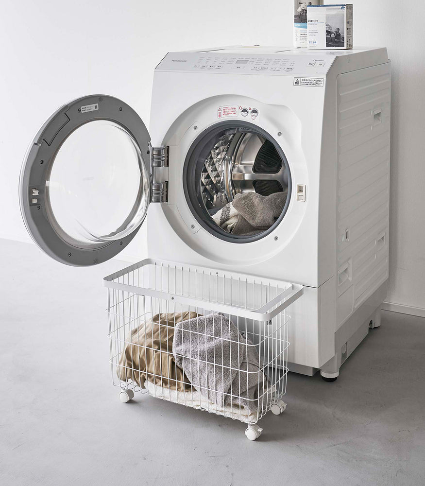 View 3 - Rolling Wire Basket by Yamazaki Home in white in front of an open dryier with several towels inside.