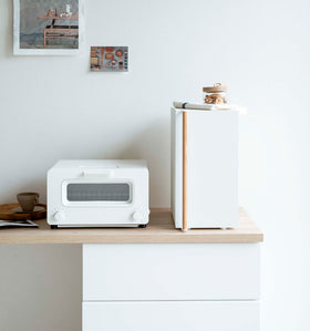 A vertical white metal breadbox, with a vertical wooden handle, is seen on a white kitchen counter next to a white microwave oven and a wooden plate with a brown coffee cup and matching saucer on it. On top of the breadbox is a folded towel and glass container of snacks. Behind the counter is a white wall with two different sized postcards positioned diagonally from each other. view 8