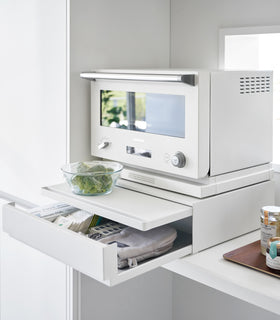 A diagonal view of the Countertop Drawer with Pull-Out Shelf by Yamazaki Home in white with both the drawer and shelf pulled out. The drawer contains food wraps, tea items, and an oven mitt among other items. A countainer of food is set on the pulled out shelf. view 4