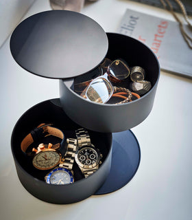 An angled bird’s-eye view of a two-tier black swivel accessory holder placed on a white dresser. The accessory holder’s tiers and lid are swiveled opened so the inside contents can be seen. In the first tier are a pair of reading glasses two silver statement rings, and an assortment of corded bracelets. In the second tier are three watches of different styles. Both compartments are lined with a felt liner. view 13
