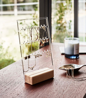 A clear acrylic translucent earring holder with a rectangular light-colored wood base are displayed on a dark wood dresser. The acrylic holder has upward pointed hooks and slots placed in an interchangeable pattern. Hanging from the hooks are chained necklaces, and in the slots are various earrings. On the surface of the dresser, in front of the earring holder, is a leaf-shaped decorative catch-all plate with necklaces strewn inside. view 3