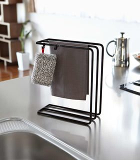 Front view of black Dish Towel Hanger holding cloth and sponge in kitchen by Yamazaki Home. view 6