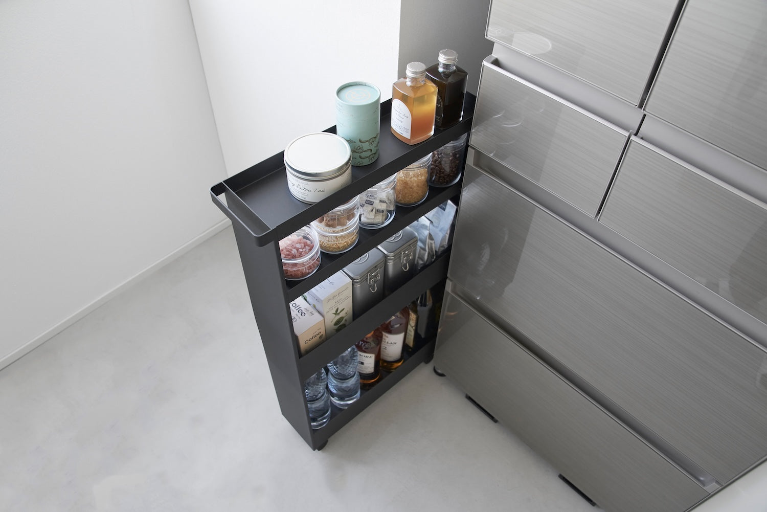 Black slim rolling wagon from yamazaki home adding storage between a fridge and the wall in a modern kitchen. It holds various pantry items like teas, spices, and oils. 