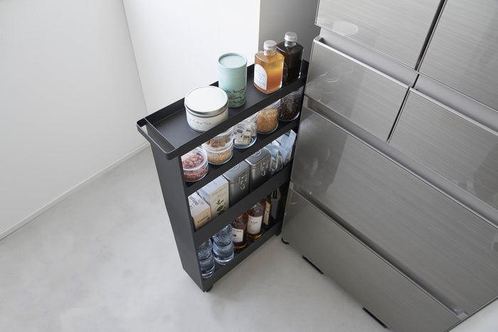 Black slim rolling wagon from yamazaki home adding storage between a fridge and the wall in a modern kitchen. It holds various pantry items like teas, spices, and oils. 