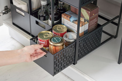 Three Two-Tier Cabinet Storage Baskets shown in a row used in a lower cabinet with cans and various dry goods. Hand pulling out one of the shelves. 