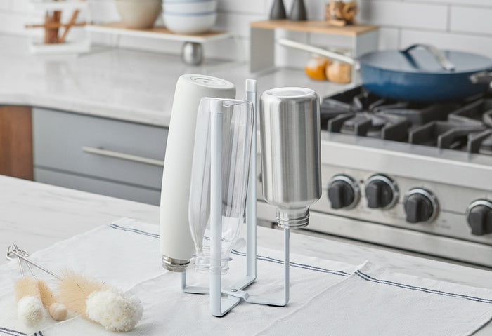 Collapsible bottle dryer in white steel, drying three water bottles and a thermos in a modern kitchen. The collapsible bottle dryer is from Yamazaki Home.