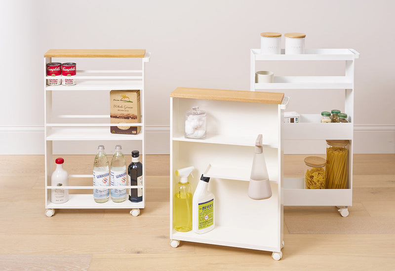 From left to right: Slim Storage Cart, Rolling Storage Cart, and the Slim Wagon all in white from Yamazaki Home. The Slim Storage Cart and Rolling Storage Cart have hard natural wood tops. All of the carts hold various household objects. 
