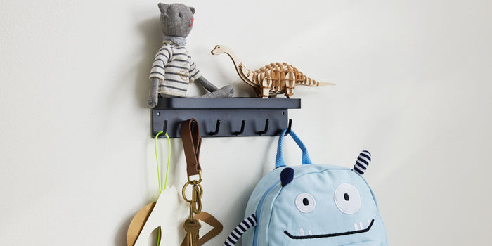 Black Magnetic Key Rack mounted on a white metal door. The upper tray holds a stuffed cat toy and a wood T-rex toy. The bottom hooks hold an arts and crafts fox mask, a set of keys, and a blue novelty child's backpack. 