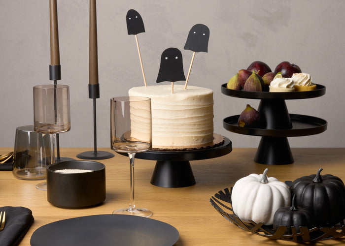 Modern halloween table set-up featuring black cakes stands and fruit bowls from Yamazaki Home. Fruit bowl holds chic black and white pumpkins. Also features small spooky treats and a cake. 