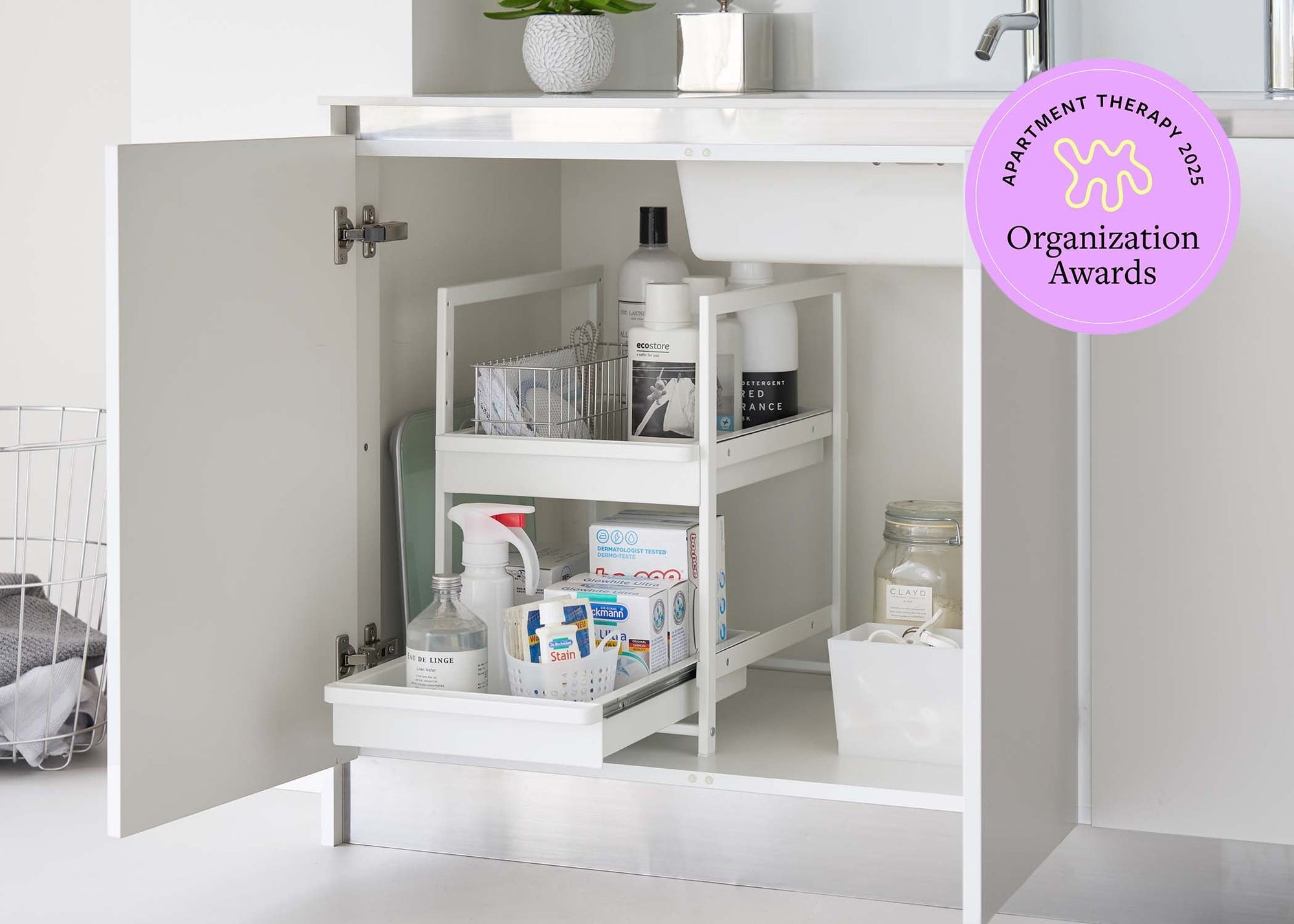 Two-Tier undershelf organizer from Yamazaki Home used in a modern bathroom holding various soaps, cleansers, and first aid supplies. Top Right corner includes a lilac 2025 Apartment Therapy Organization Awards Logo