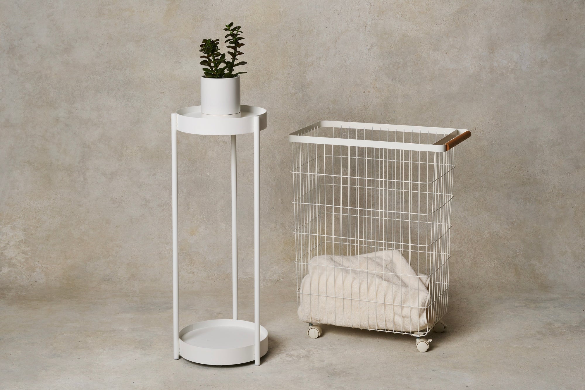 Two-tier plant stand made of white steel from Yamazaki Home next to a wire cart rolling basket from Yamazaki Home with a wood handle, holding a cozy blanket. Gift Guide for Homebodies 2025