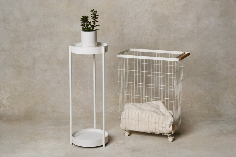 Two-tier plant stand made of white steel from Yamazaki Home next to a wire cart rolling basket from Yamazaki Home with a wood handle, holding a cozy blanket. Gift Guide for Homebodies 2025