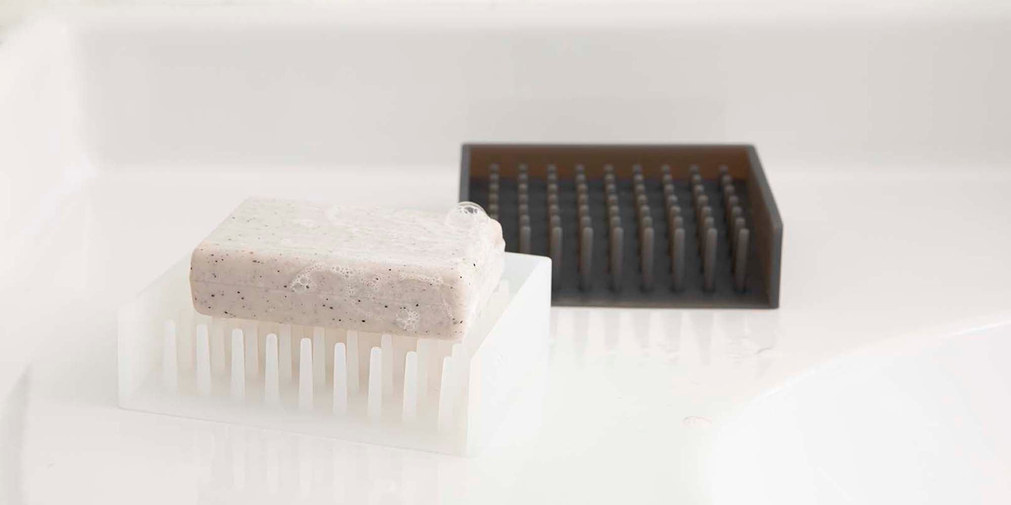 Two Self-Draining Soap Dishes. White soap dish holds bar freshly used bar of soap. Black soap dish sits behind the former. 