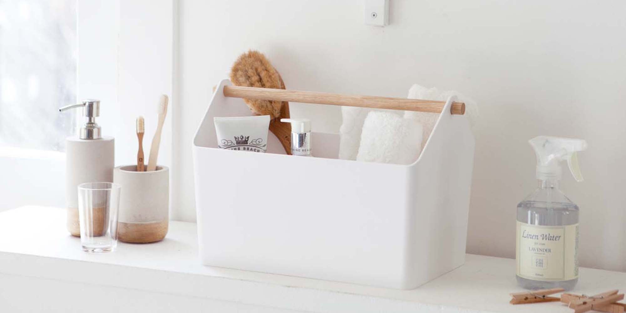 White Yamazaki Home storage caddy with wood handle holding various bathroom supplies like a dry brush, wash cloths, and serums.